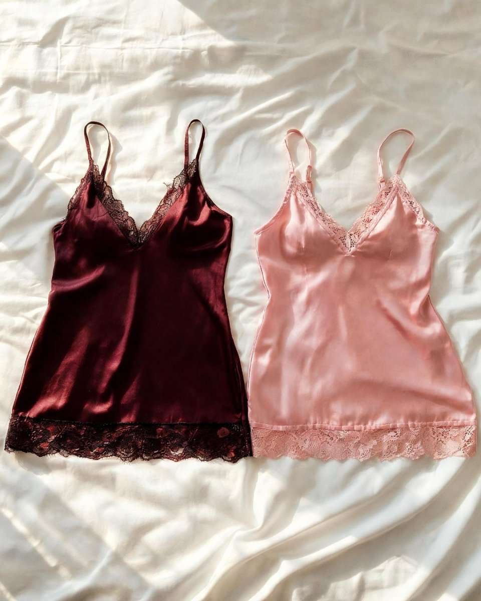 Two satin camisole chemises with lace trim, one in deep burgundy and one in light pink, laid flat on a white satin sheet.