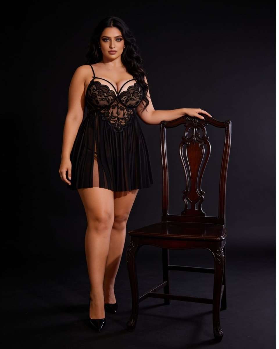A woman models a black lace and mesh mini lingerie dress with a high slit, standing next to a dark wood chair against a black studio background.