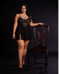A woman models a black lace and mesh mini lingerie dress with a high slit, standing next to a dark wood chair against a black studio background.