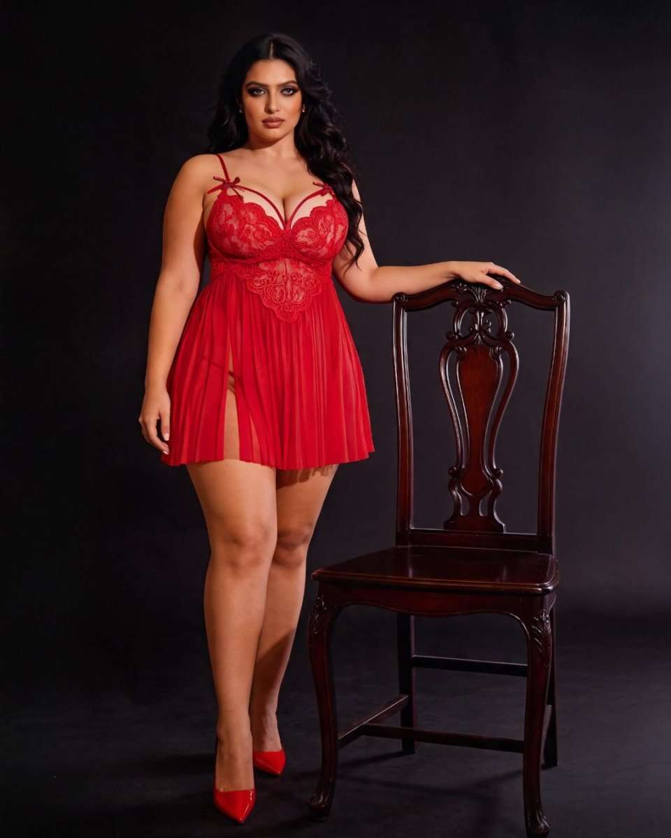A woman models a red lace and mesh babydoll lingerie dress next to a dark wooden chair against a black studio background.