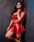 A woman posing in a shiny red two-piece outfit consisting of a sequined crop top with fringe and a matching pleated mini skirt, set against a dark textured studio wall.