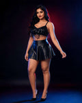 A woman modeling a black two-piece outfit consisting of a fringed spaghetti-strap crop top and a black faux-leather mini skirt, paired with black high heels, set against a dark studio background with red and blue lighting.