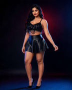 A woman modeling a black two-piece outfit consisting of a fringed spaghetti-strap crop top and a black faux-leather mini skirt, paired with black high heels, set against a dark studio background with red and blue lighting.