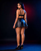 A woman stands with her back to the camera wearing a black strappy bralette and a shiny black faux-leather mini skirt, paired with black high heels, against a dark studio background with blue and red lighting.
