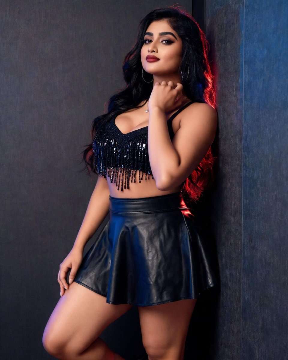 A woman posing in a black sequined fringe crop top and a black faux-leather mini skirt against a dark textured wall.