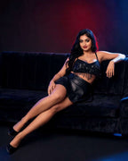 A woman reclining on a black velvet couch wearing a black fringe-embellished crop top and matching mini skirt against a dark studio background with red and blue lighting.