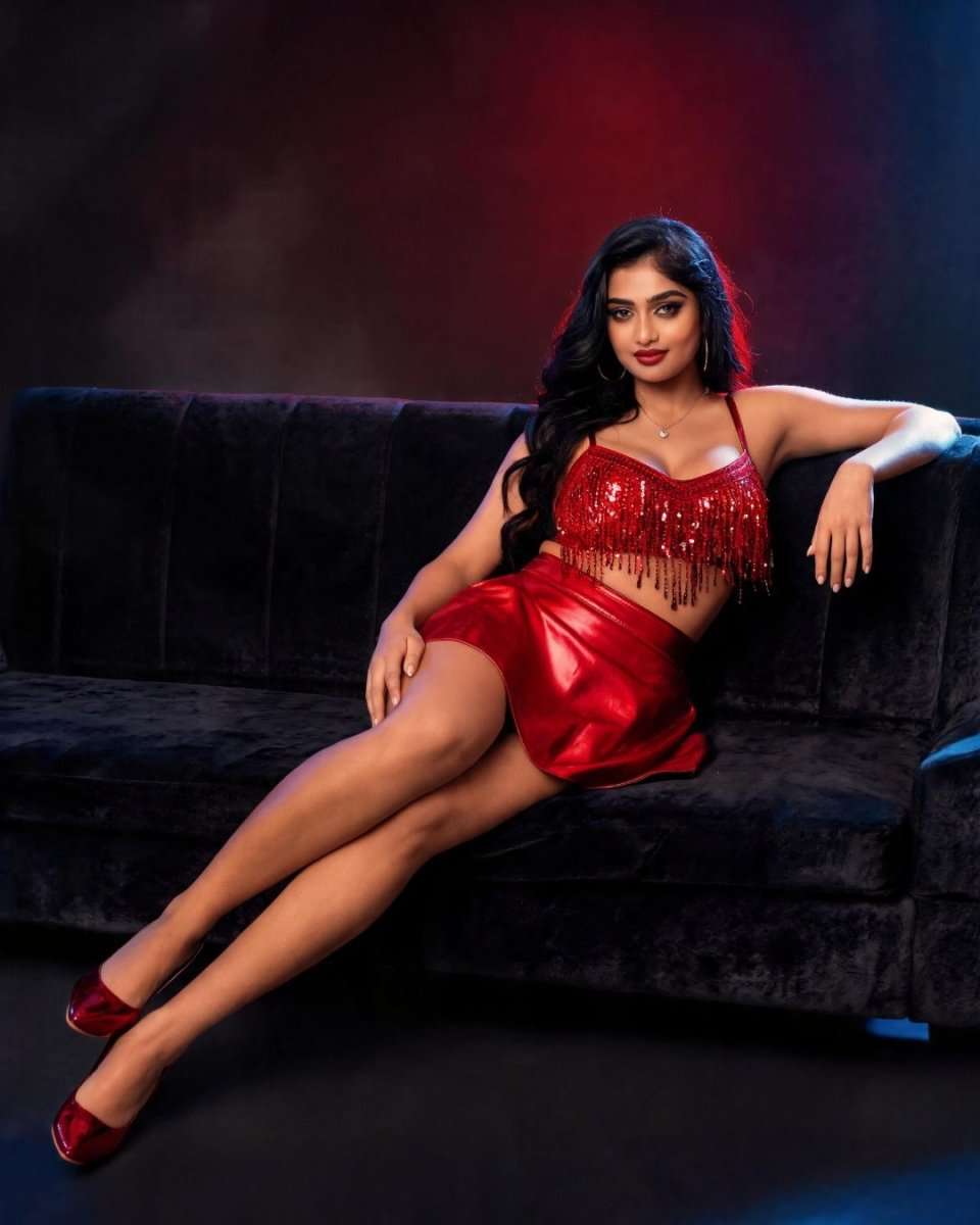 A woman reclining on a dark velvet couch wearing a shiny red two-piece outfit with a fringed crop top and matching mini skirt, posed in a dimly lit studio with red and blue lighting.