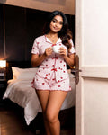 A woman stands in a doorway wearing a pink and white striped pajama set with red heart patterns, holding a white mug in a bedroom setting.