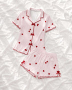 A two-piece women's pajama set consisting of a short-sleeve button-front top and matching shorts, laid flat on a white textured fabric background. The pajamas feature a light pink and white vertical stripe pattern, red heart motifs, and red piping details including bow closures on the front and ties on the shorts.