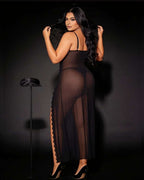 A woman wearing a sheer black maxi dress with a high side slit, posing from behind against a dark studio background. She is wearing black high heels, and a small black hat sits on a stand to her left.