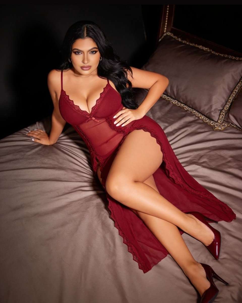 A woman reclines on a bed wearing a sheer burgundy lace-trim lingerie dress with a high slit and long flowing overlay, paired with matching pointed heels.