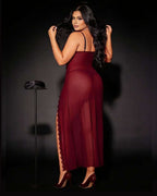 A woman modeling a sheer burgundy maxi dress with a high side slit, photographed against a dark studio background. She is wearing burgundy high heels, and a matching small hat sits on a stand to the left.