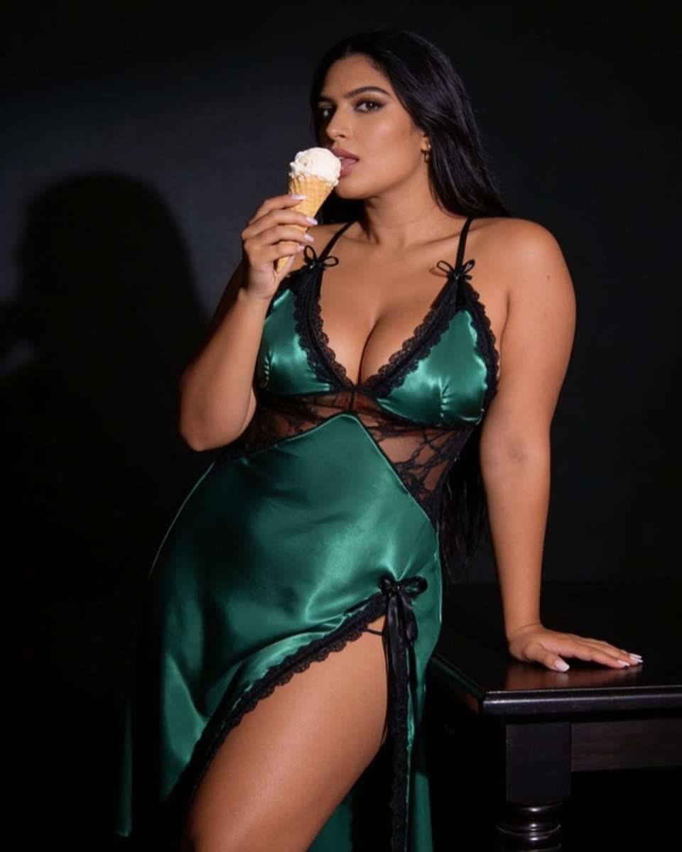 A woman wearing a satin green lingerie dress with black lace trim and sheer black mesh panels, holding an ice cream cone against a dark studio background.