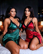 Two women posing on a bed wearing satin lingerie chemises in green and red with black lace trim and black thong undergarments in a dimly lit bedroom setting.
