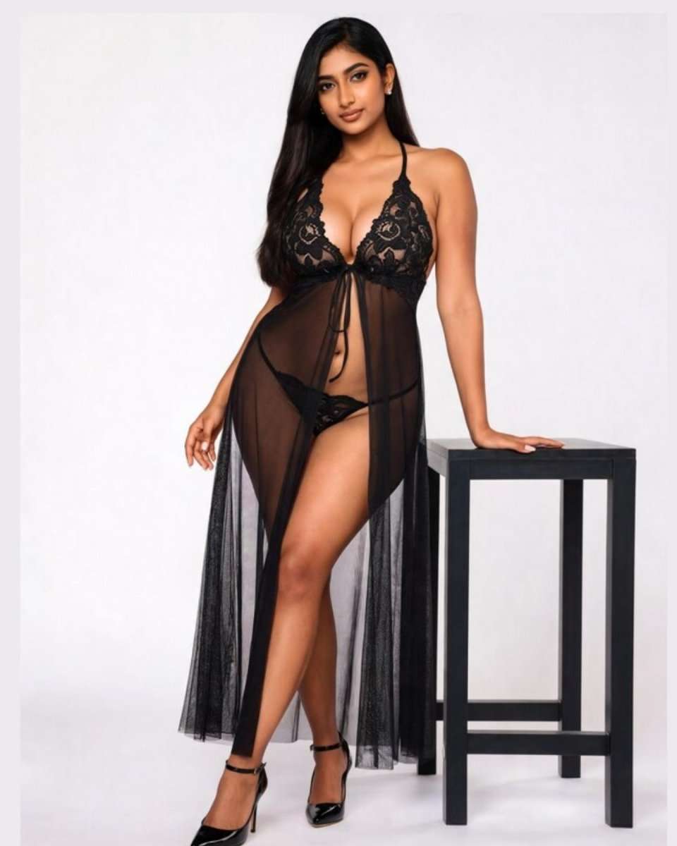 A woman models a black sheer lace babydoll lingerie chemise with a high leg slit, standing next to a black stool against a white studio background.