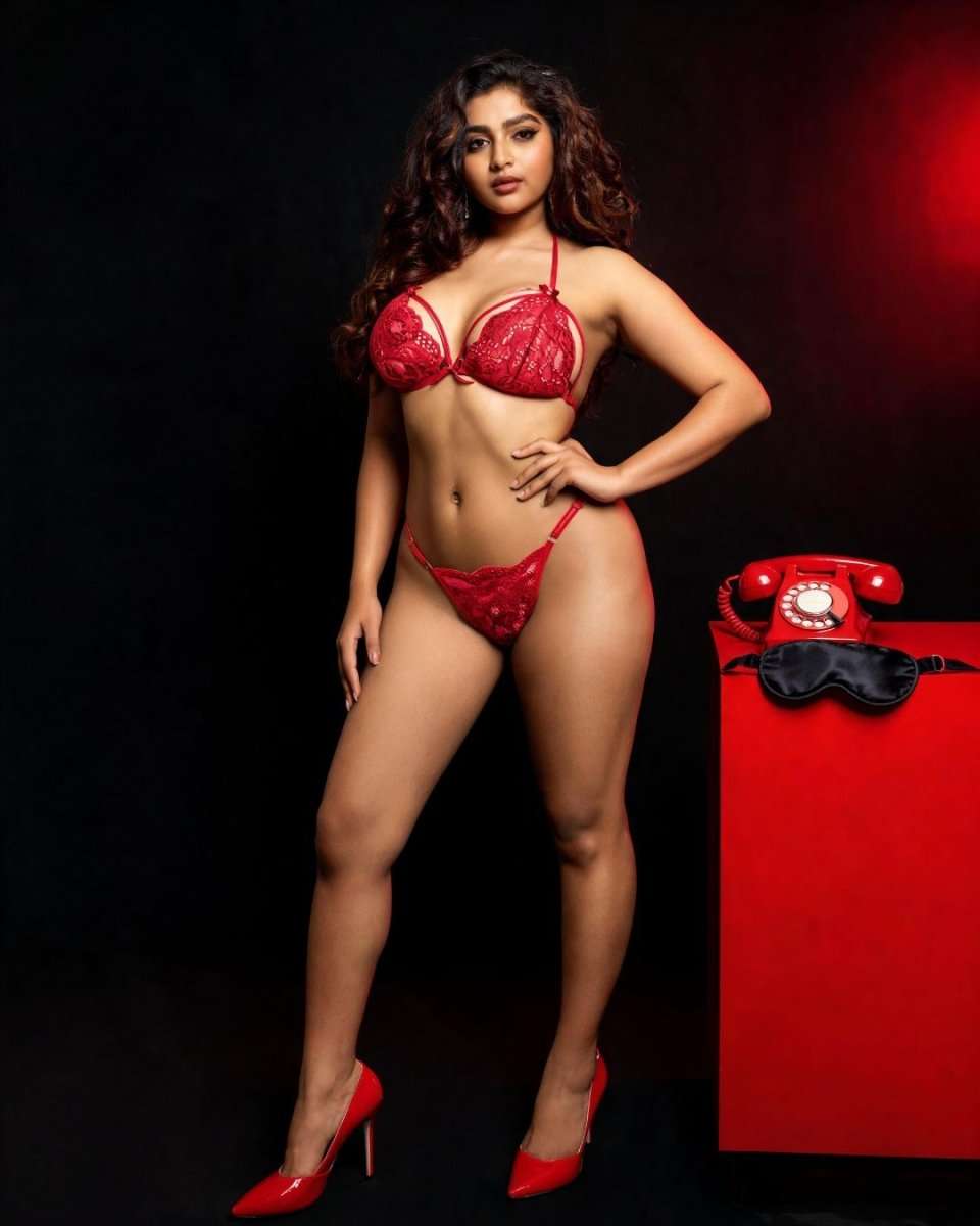 A woman modeling a red lace lingerie set consisting of a bra and matching thong, standing against a dark studio background. A red vintage rotary phone and a black eye mask rest on a red pedestal to her right.