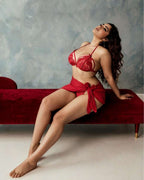 A woman poses on a red velvet bench wearing a red lace lingerie set with a matching red fabric wrap, set against a textured gray wall.