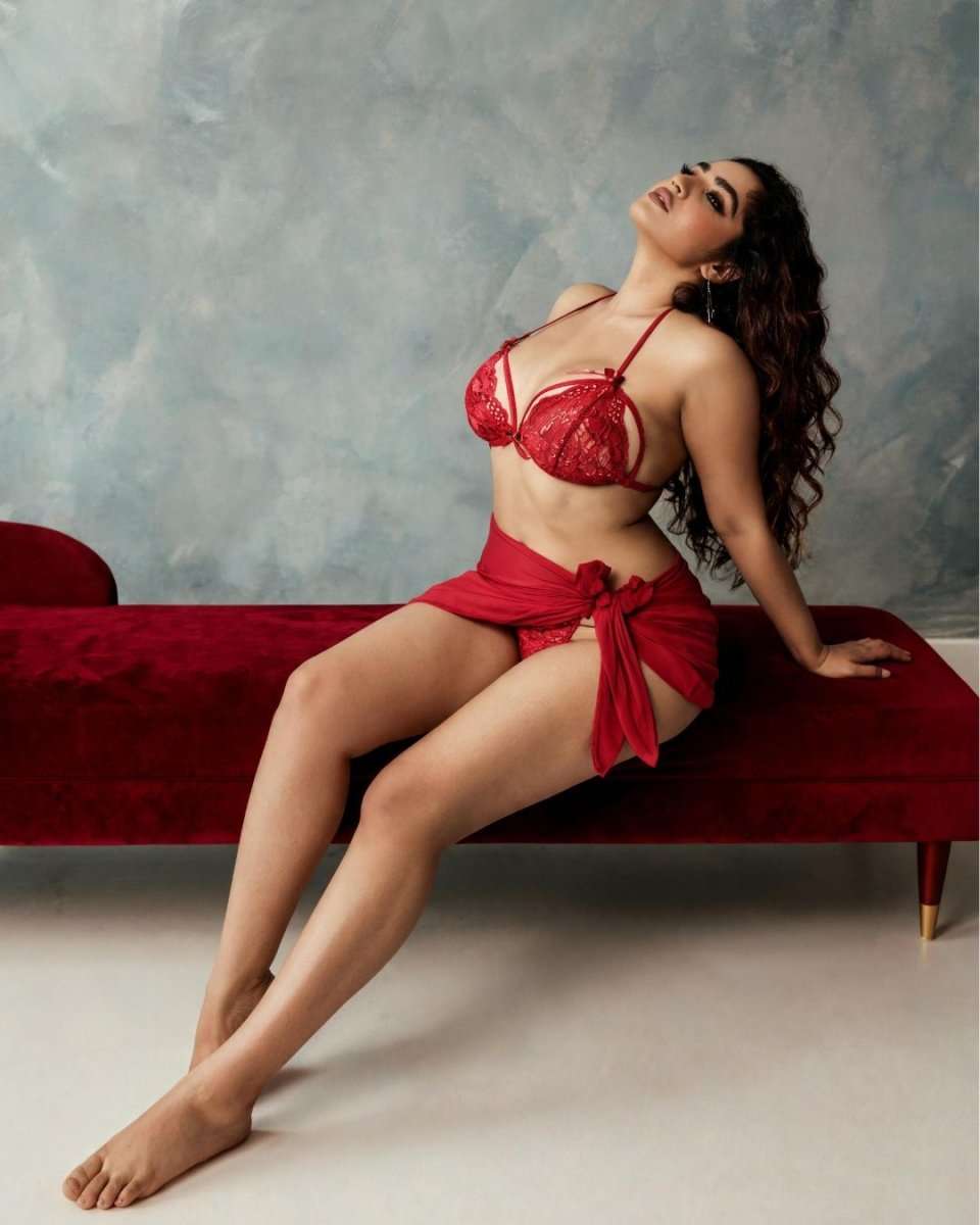 A woman poses on a red velvet bench wearing a red lace lingerie set with a matching red fabric wrap, set against a textured gray wall.