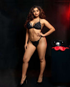 A woman modeling a black lace lingerie set consisting of a bra and matching thong, standing against a dark studio background with a red light accent. A vintage black rotary phone and a red satin bow are displayed on a pedestal to her right.