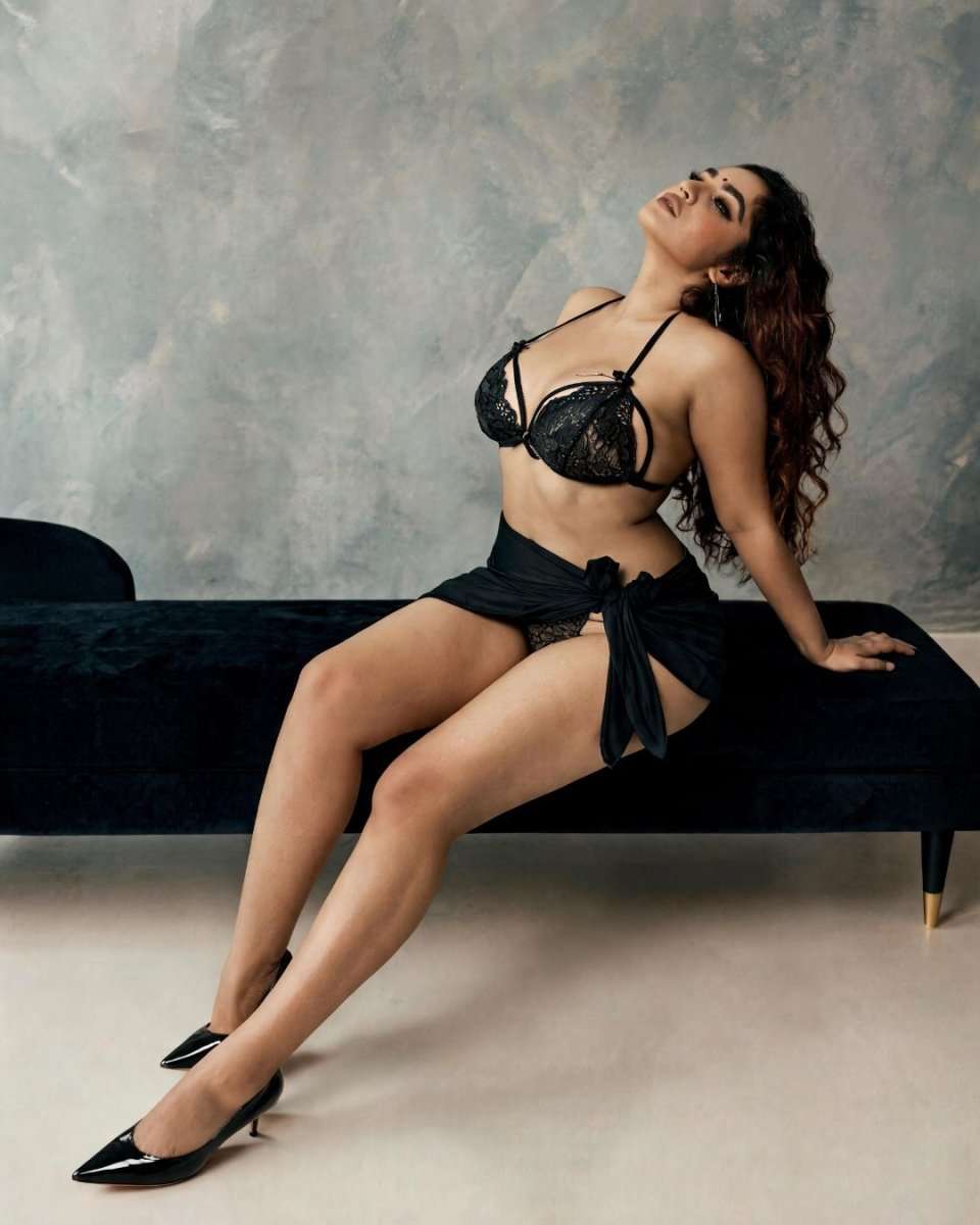 A woman poses on a black velvet bench wearing a black lace lingerie set with a matching tied skirt and black pointed-toe heels, set against a textured gray wall.