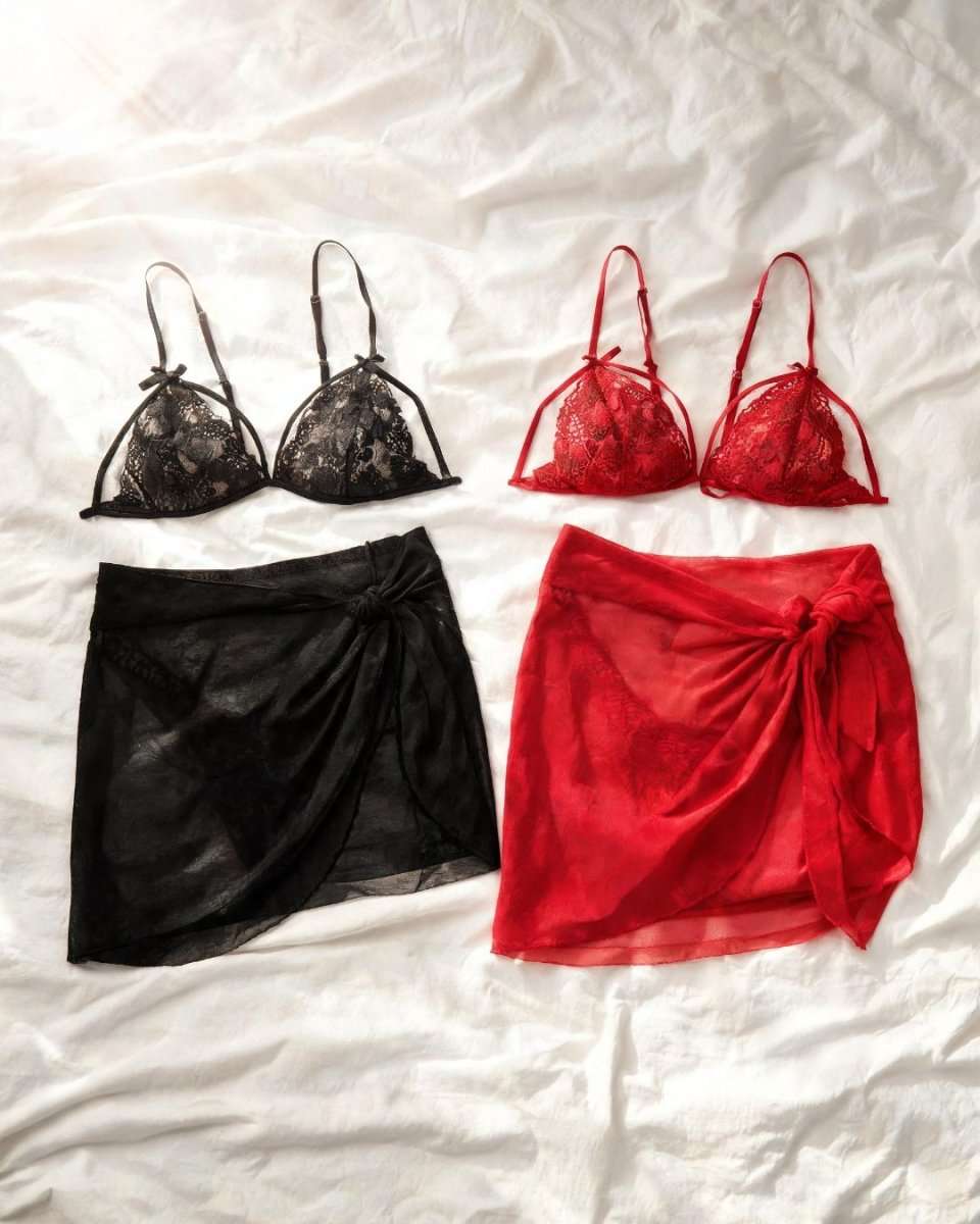 Two matching lingerie sets laid flat on white fabric: a black lace triangle bra with a black sheer wrap skirt, and a red lace triangle bra with a red sheer wrap skirt.