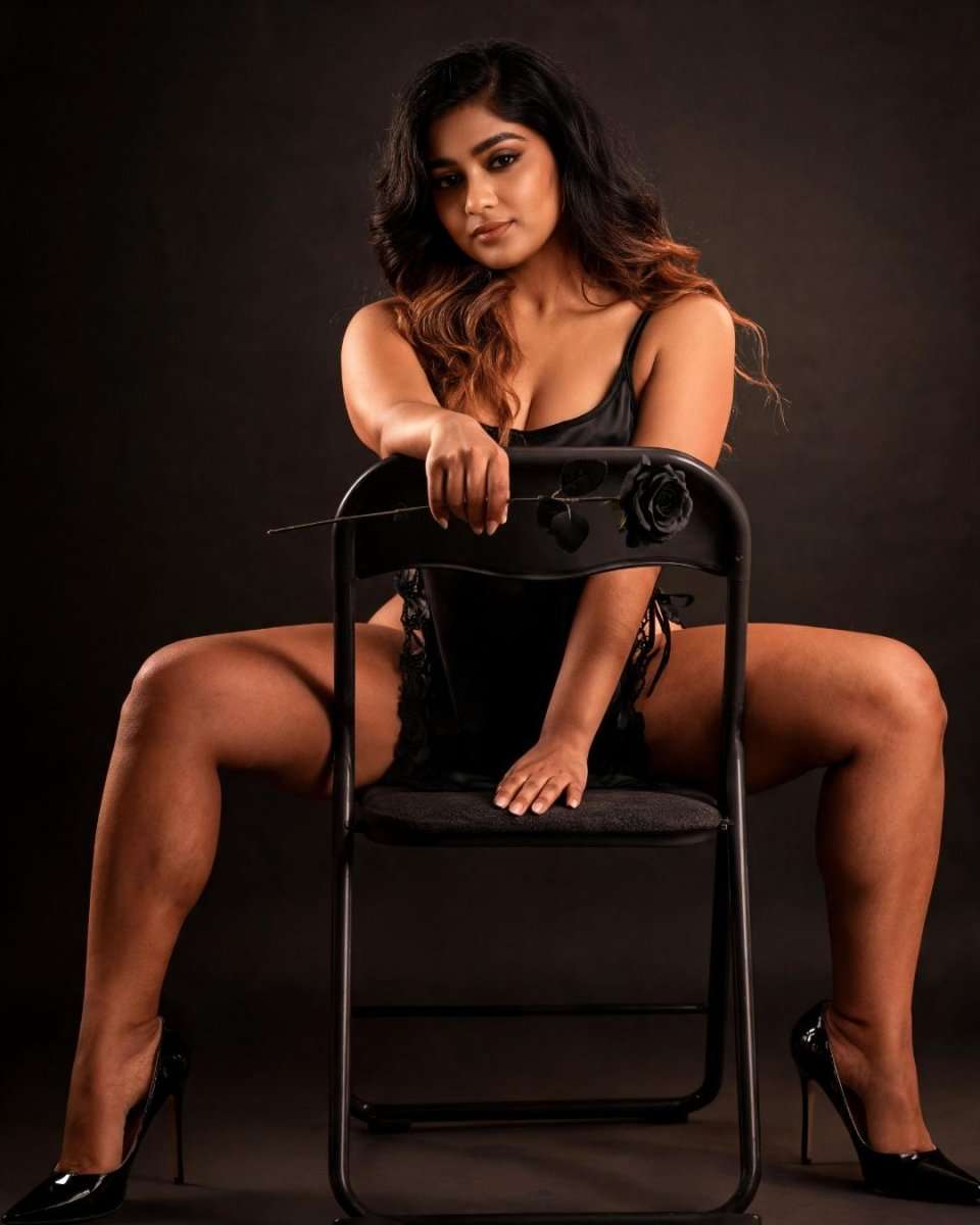 A woman posing on a black folding chair against a dark brown studio background, wearing a black lingerie bodysuit and black high heels.