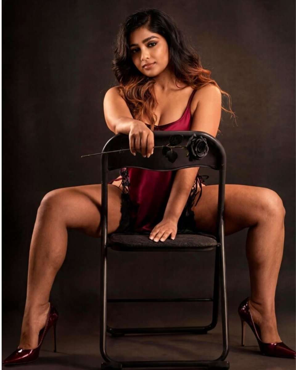A woman posing on a black folding chair in a dark studio setting, wearing a deep red lingerie dress and matching red high heels.