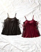 Two satin camisole sleepwear tops laid flat on a white wrinkled sheet. The left top is black with lace-up side details, and the right top is deep burgundy with matching lace-up side details.