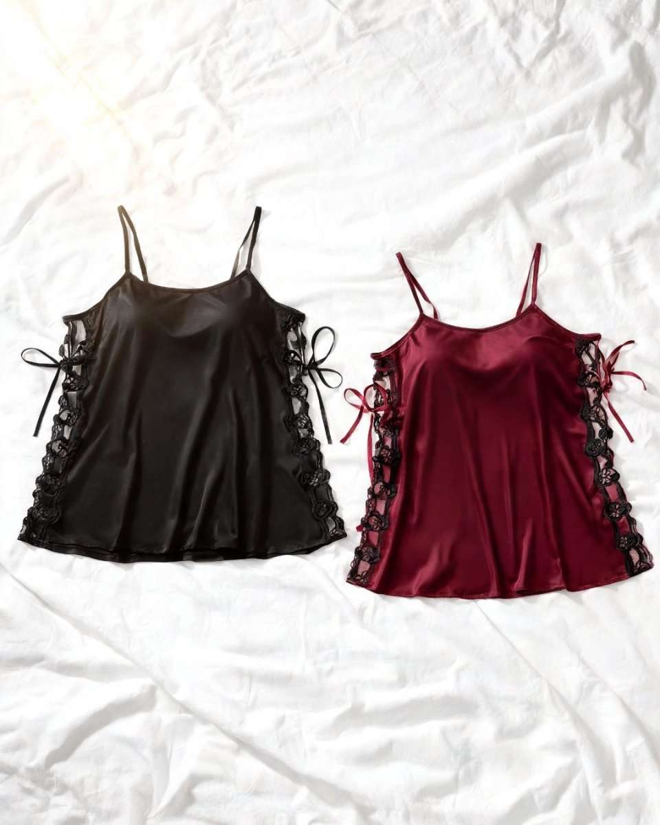 Two satin camisole sleepwear tops laid flat on a white wrinkled sheet. The left top is black with lace-up side details, and the right top is deep burgundy with matching lace-up side details.