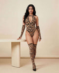 A woman modeling a leopard-print halter bodysuit with matching thigh-high stockings and black heels, standing beside a minimalist beige table against a neutral beige studio backdrop.