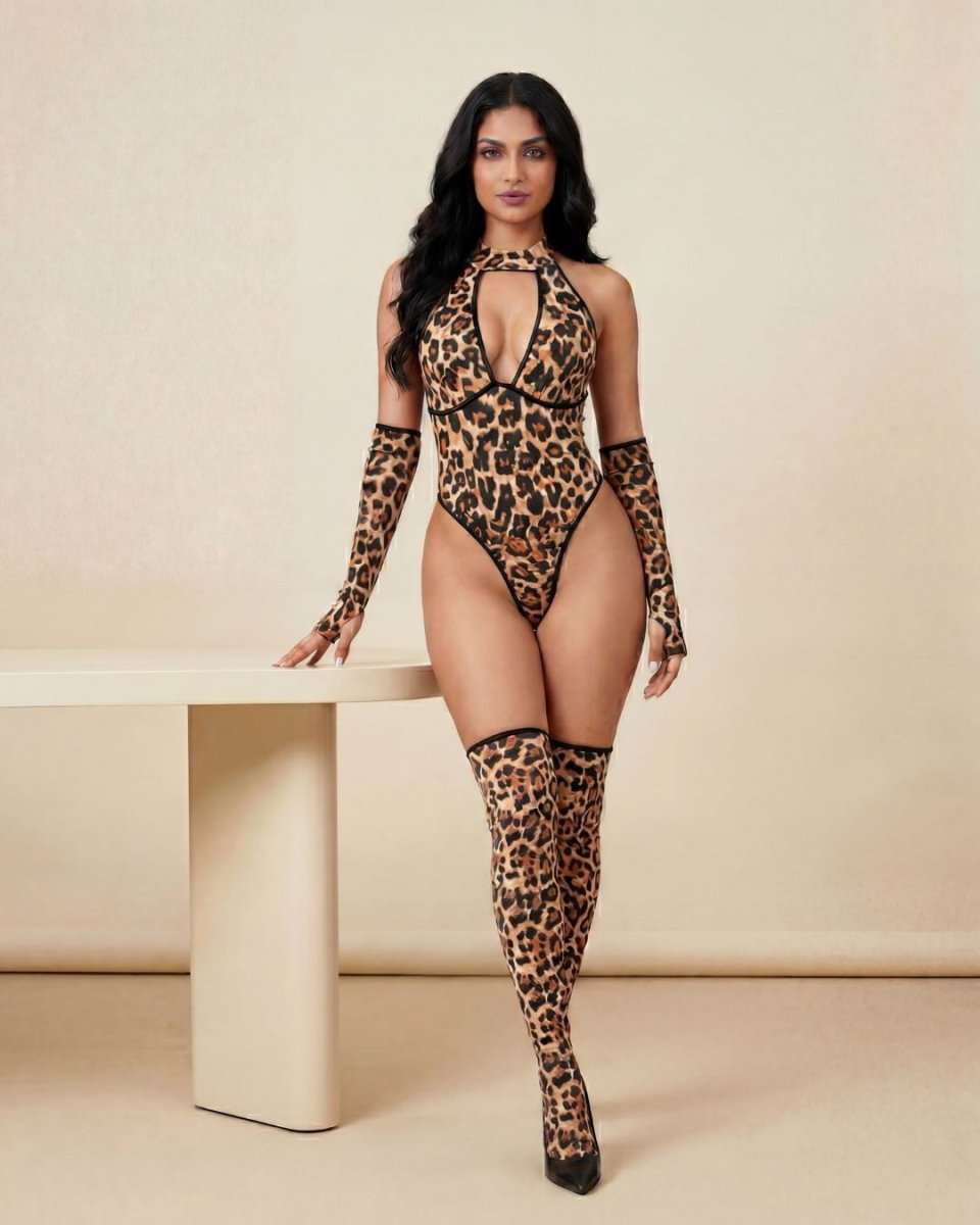 A woman modeling a leopard-print halter bodysuit with matching thigh-high stockings and black heels, standing beside a minimalist beige table against a neutral beige studio backdrop.