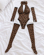 A leopard-print lingerie set laid flat on white bedding, including a halter bodysuit with a keyhole cutout, matching thigh-high stockings, and two long arm sleeves.